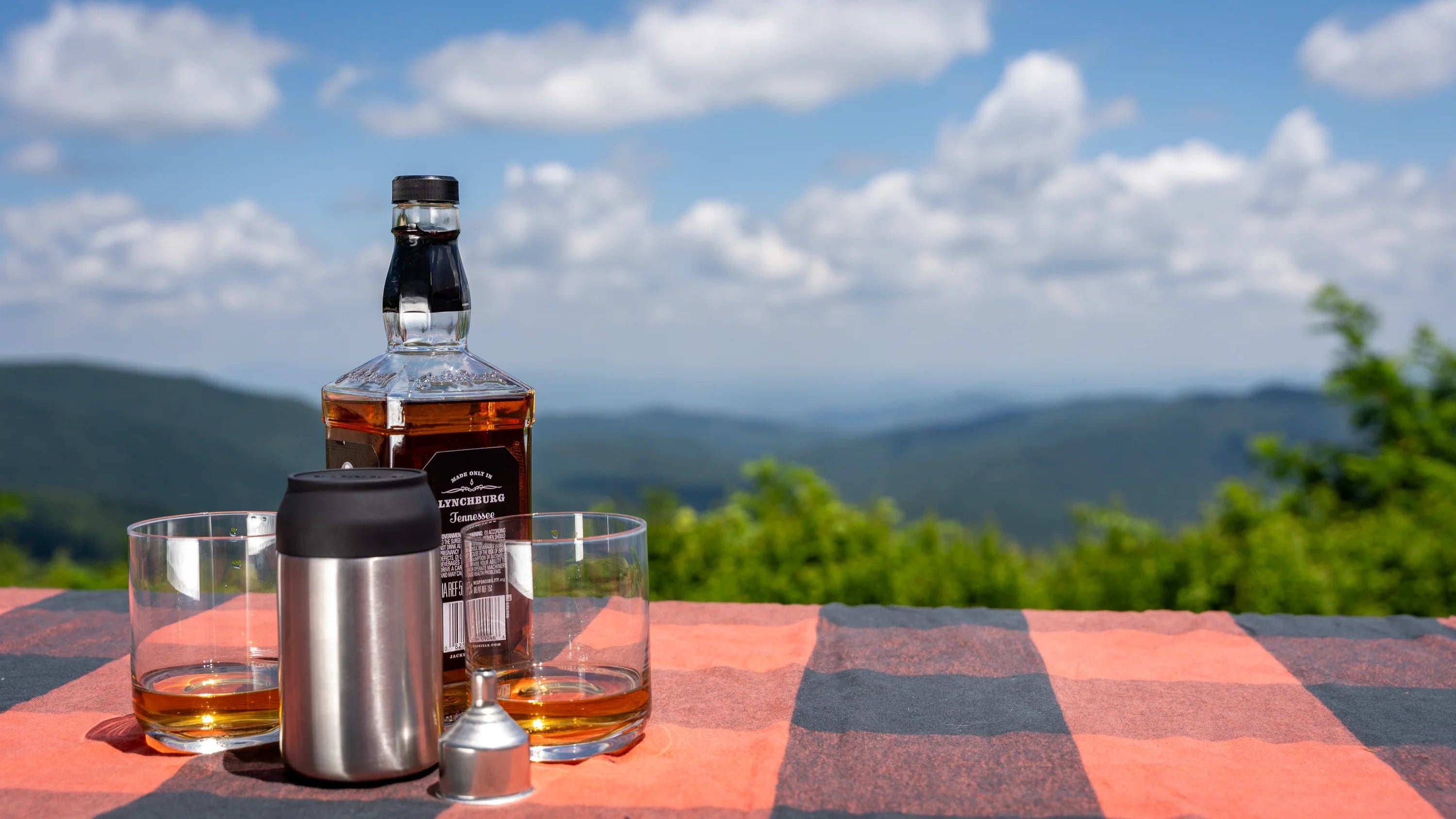 Bottle of whiskey with glasses on a checkered tablecloth against a scenic mountain backdrop